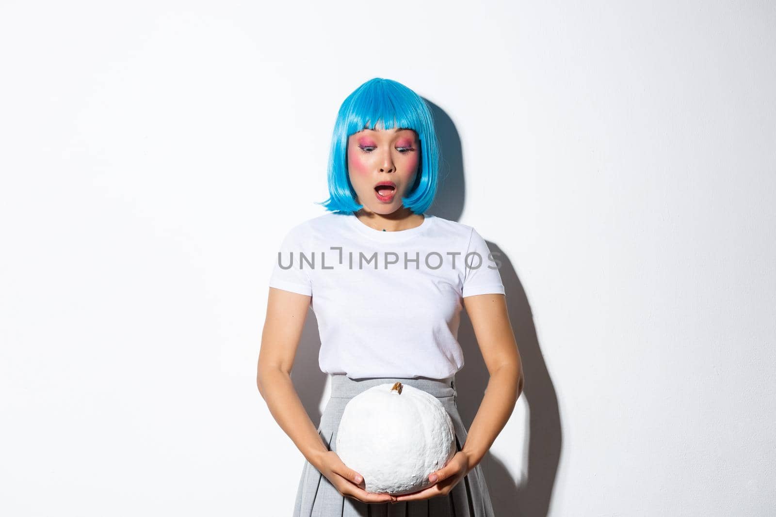 Image of surprised asian girl in blue wig, gasping and looking amazed at white pumpkin, celebrating halloween, standing over white background.