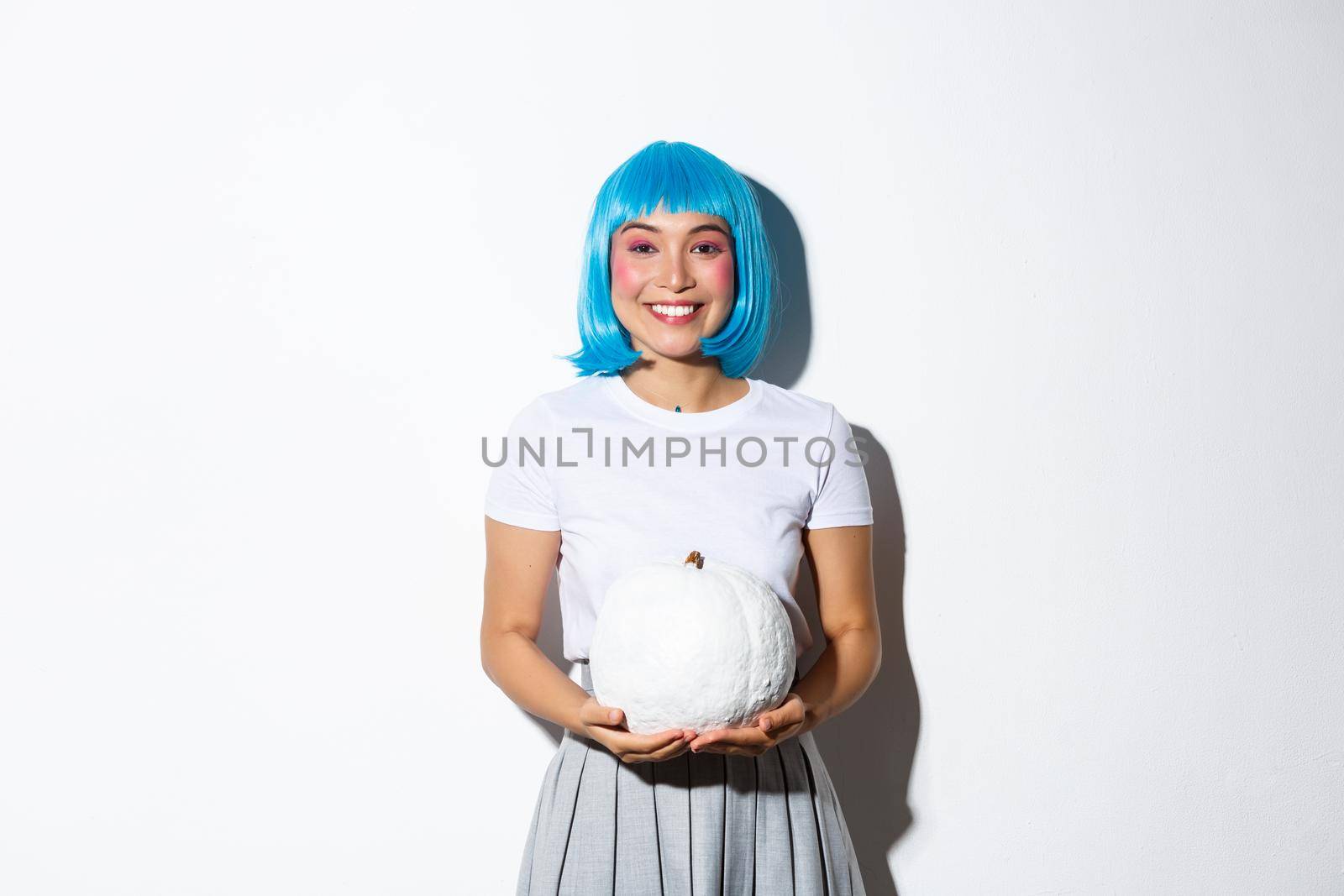 Beautiful asian girl in blue wig holding white pumpkin and smiling happy, celebrating halloween, standing over white background.