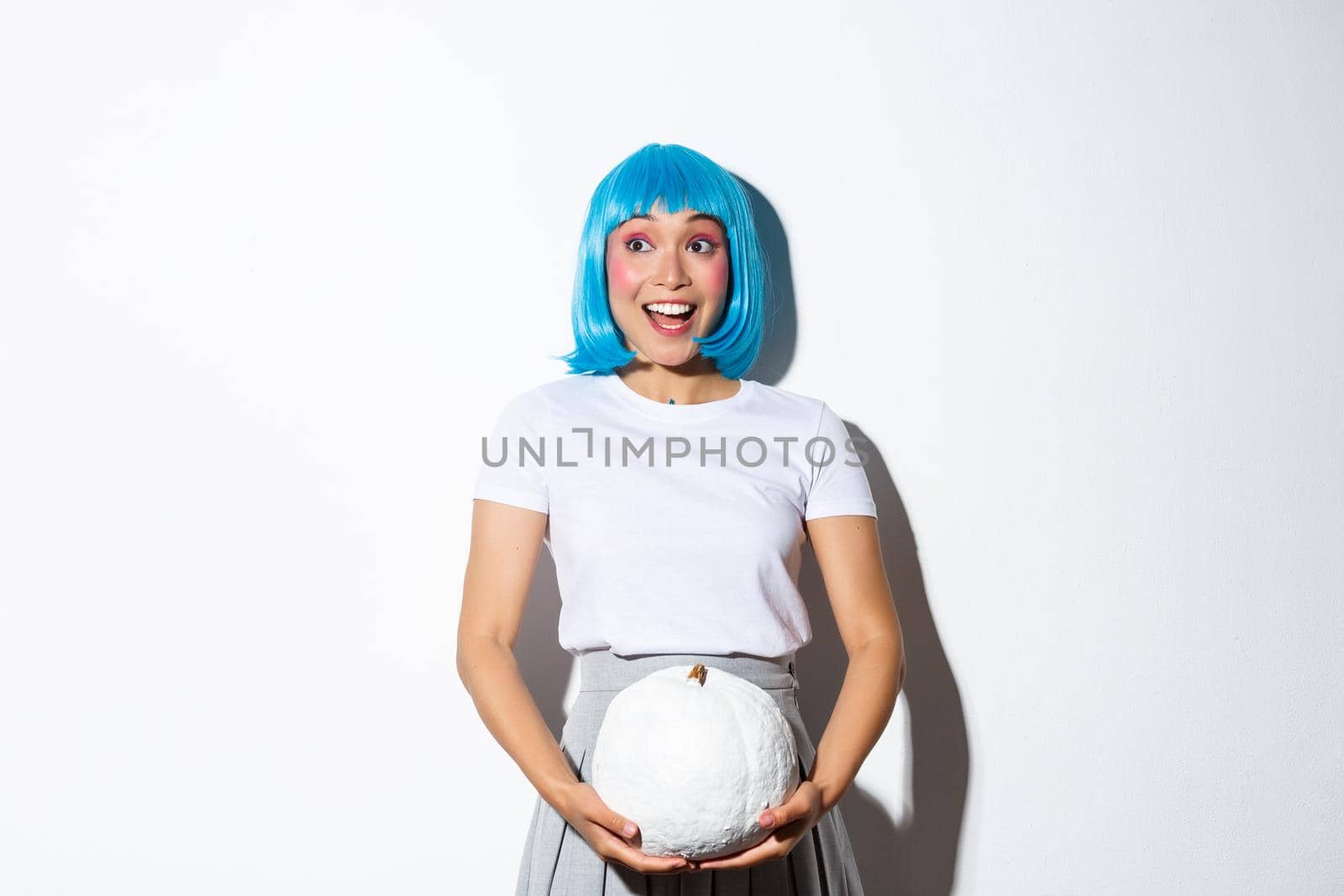 Image of excited cute asian girl looking at upper left corner banner about halloween, holding white pumpkin, wearing blue wig for party.