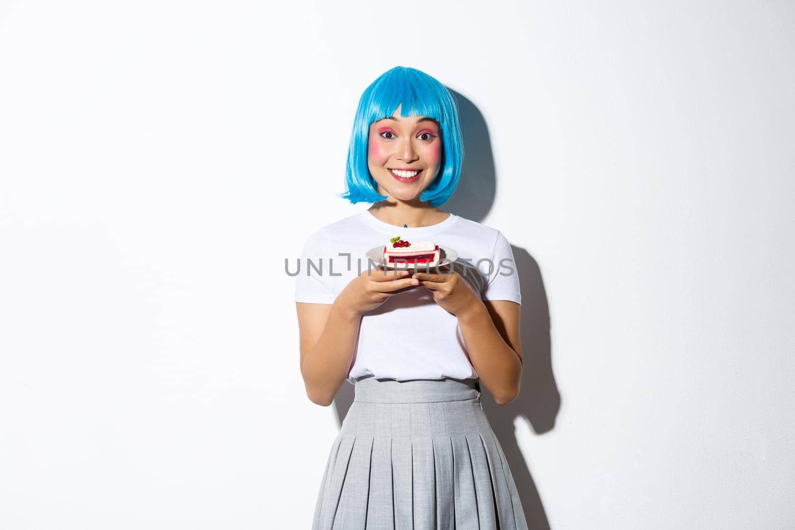 Image of cheerful cute asian girl in blue short wig and schoolgirl costume, smiling happy, holding piece of cake, standing over white background.