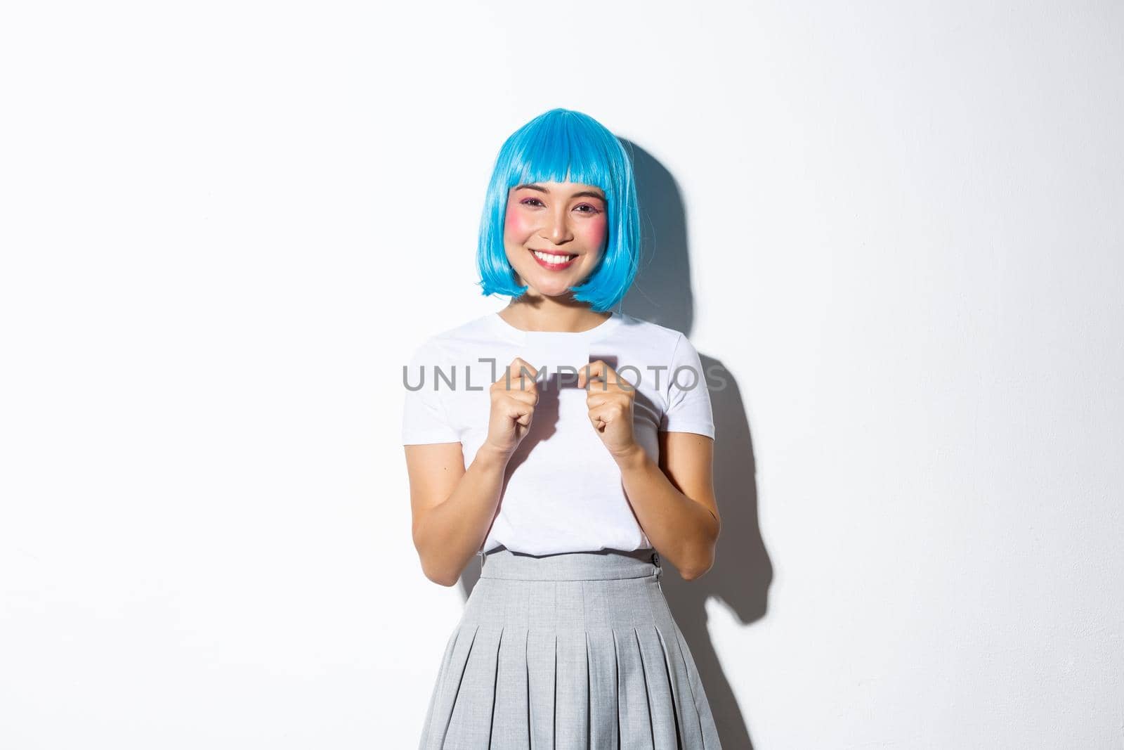 Smiling pretty asian girl showing credit card, wearing blue wig for halloween party, standing over white background.