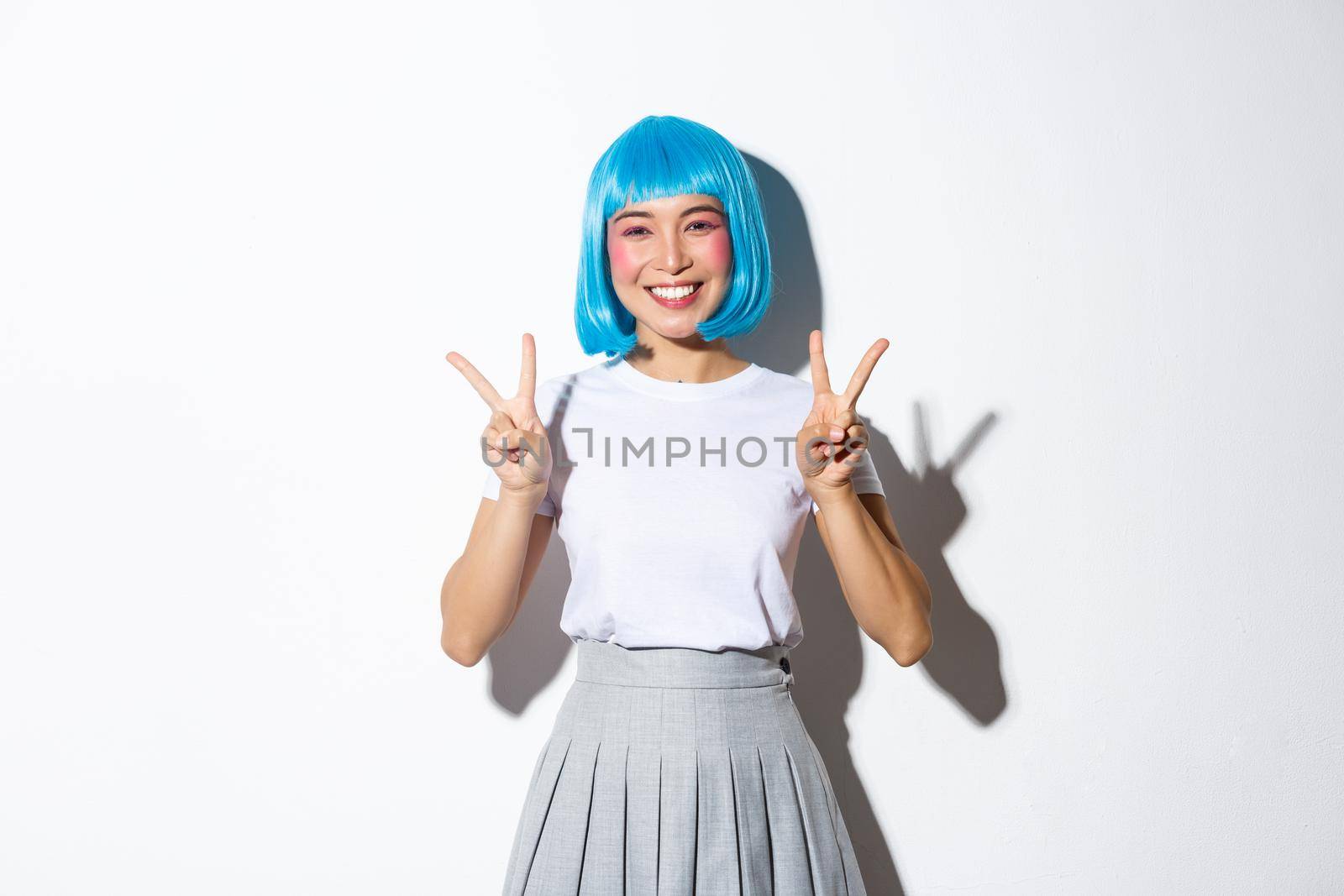 Cheerful asian girl in blue party wig, looking cute in halloween costume and showing peace gestures, standing kawaii over white background.