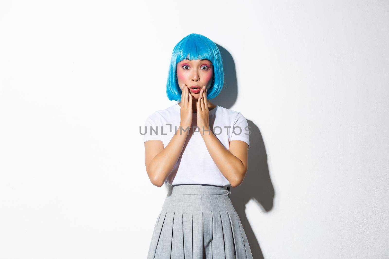 Surprised asian kawaii girl in blue short wig, gasping amazed, holding hands near mouth and looking at camera, wearing halloween costume, standing over white background.