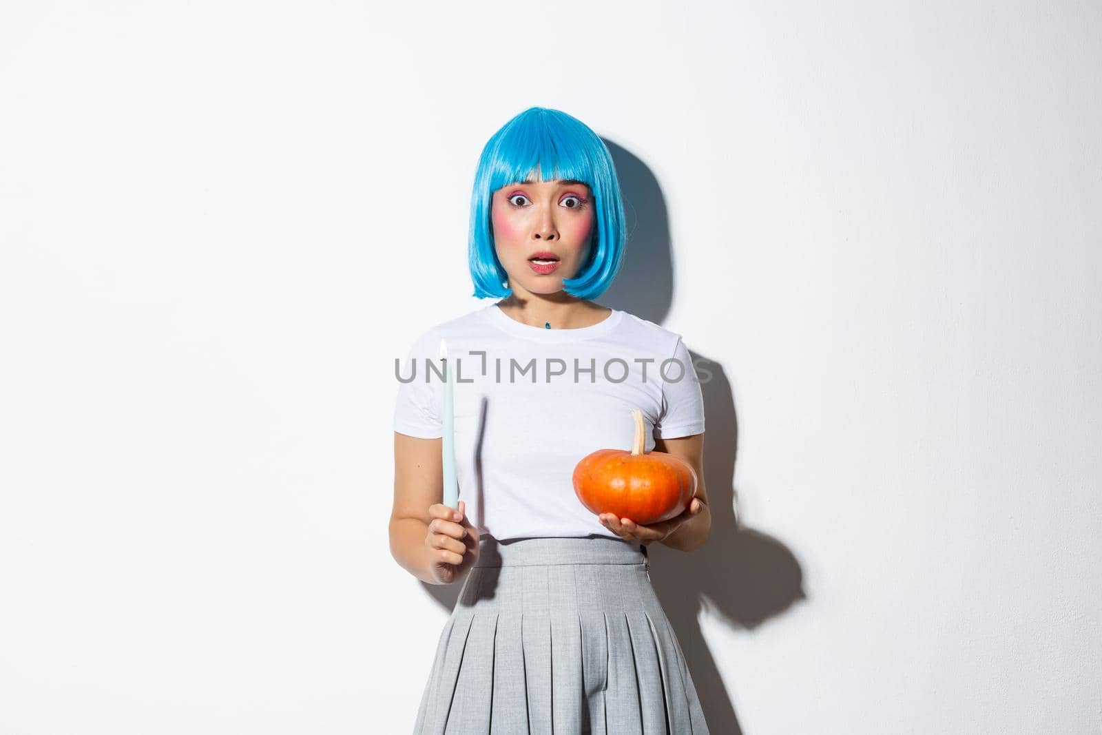 Concept of halloween. Image of scared asian girl in blue wig looking nervous and frightened, holding candle and pumpkin by Benzoix