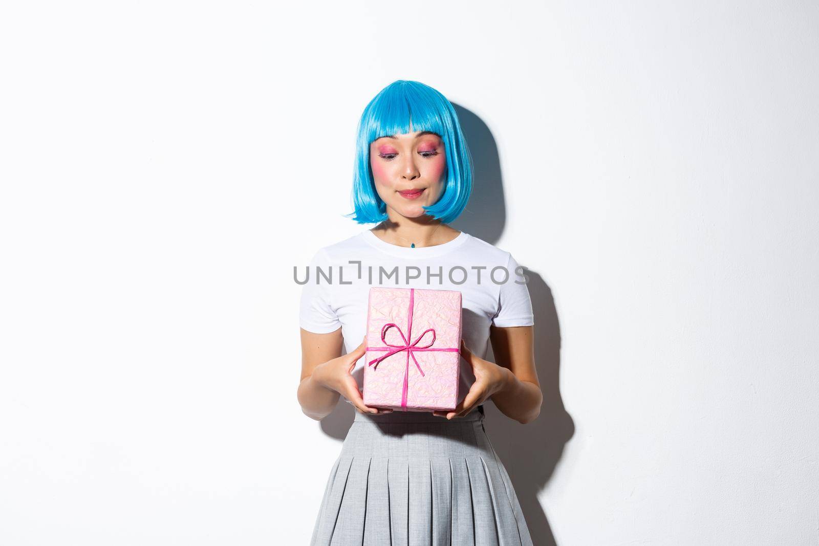 Image of cute b-day girl in blue party wig, looking curious at giftbox, standing over white background.