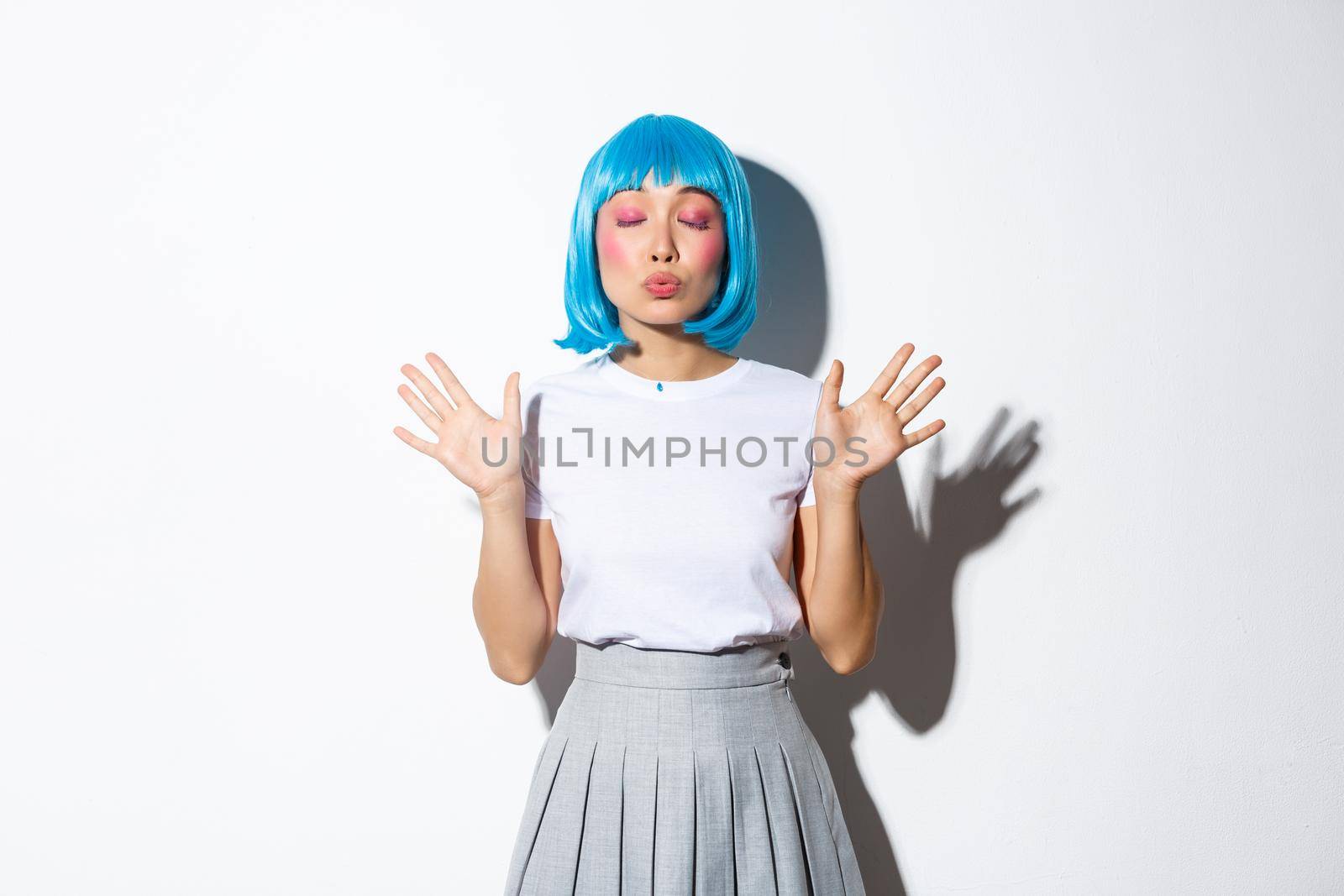 Portrait of cute asian girl in blue party wig, close eyes and pouting for kiss, standing over white background.