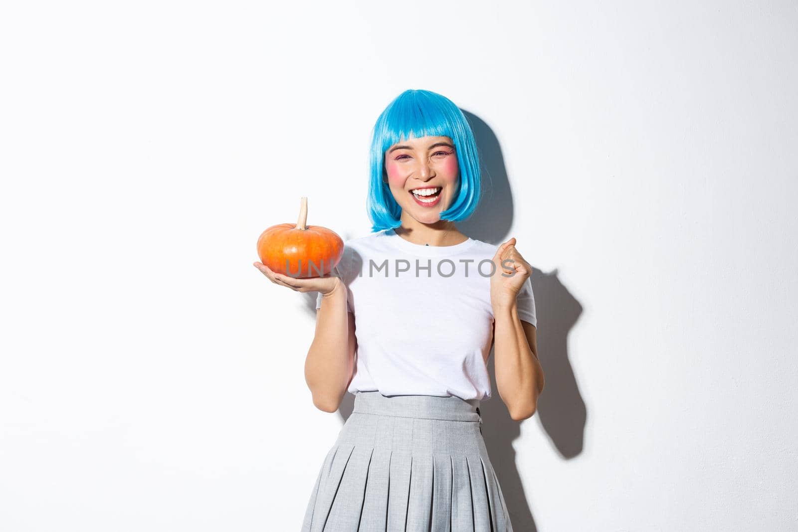 Cheerful cute asian girl celebrating halloween, wearing party costume and wig, holding small pumpkin, standing over white background.