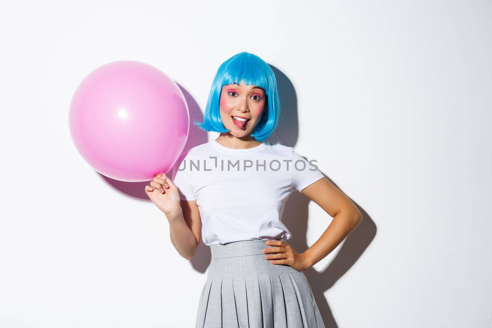 Image of silly party girl in blue wig celebrating holiday, holding pink balloon and showing tongue, standing over white backgound.