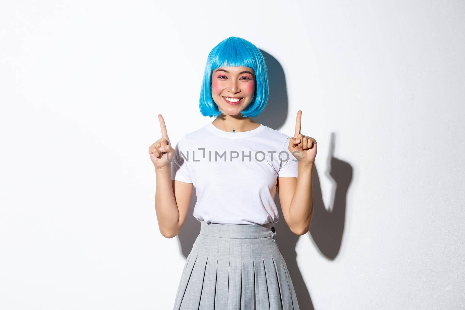 Cheerful asian woman in anime style outfit, wearing blue wig and smiling happy, pointing fingers up, showing advertisement, standing over white background.