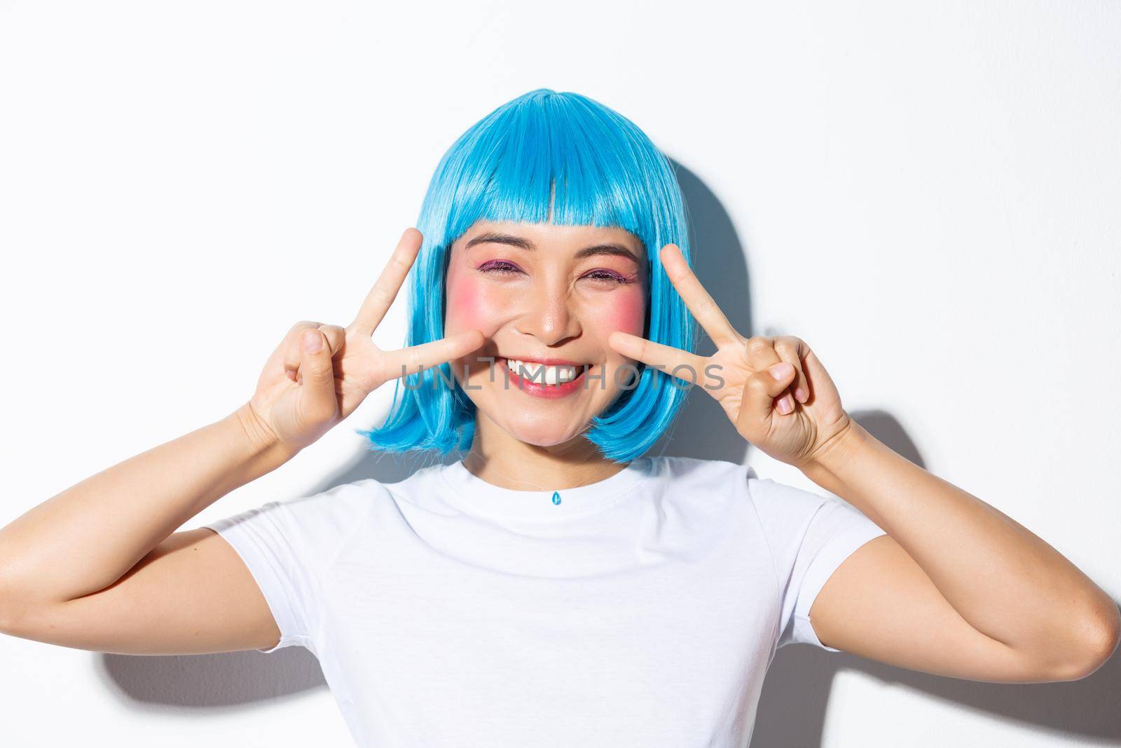 Image of cute asian woman in blue wig showing kawaii peace gesture and smiling, celebrating halloween, standing over white background by Benzoix