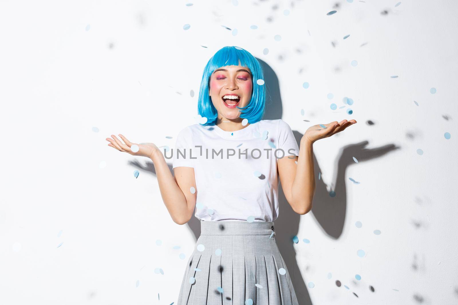 Portrait of happy asian girl in blue wig celebrating halloween, throwing confetti in the air, standing over white background.