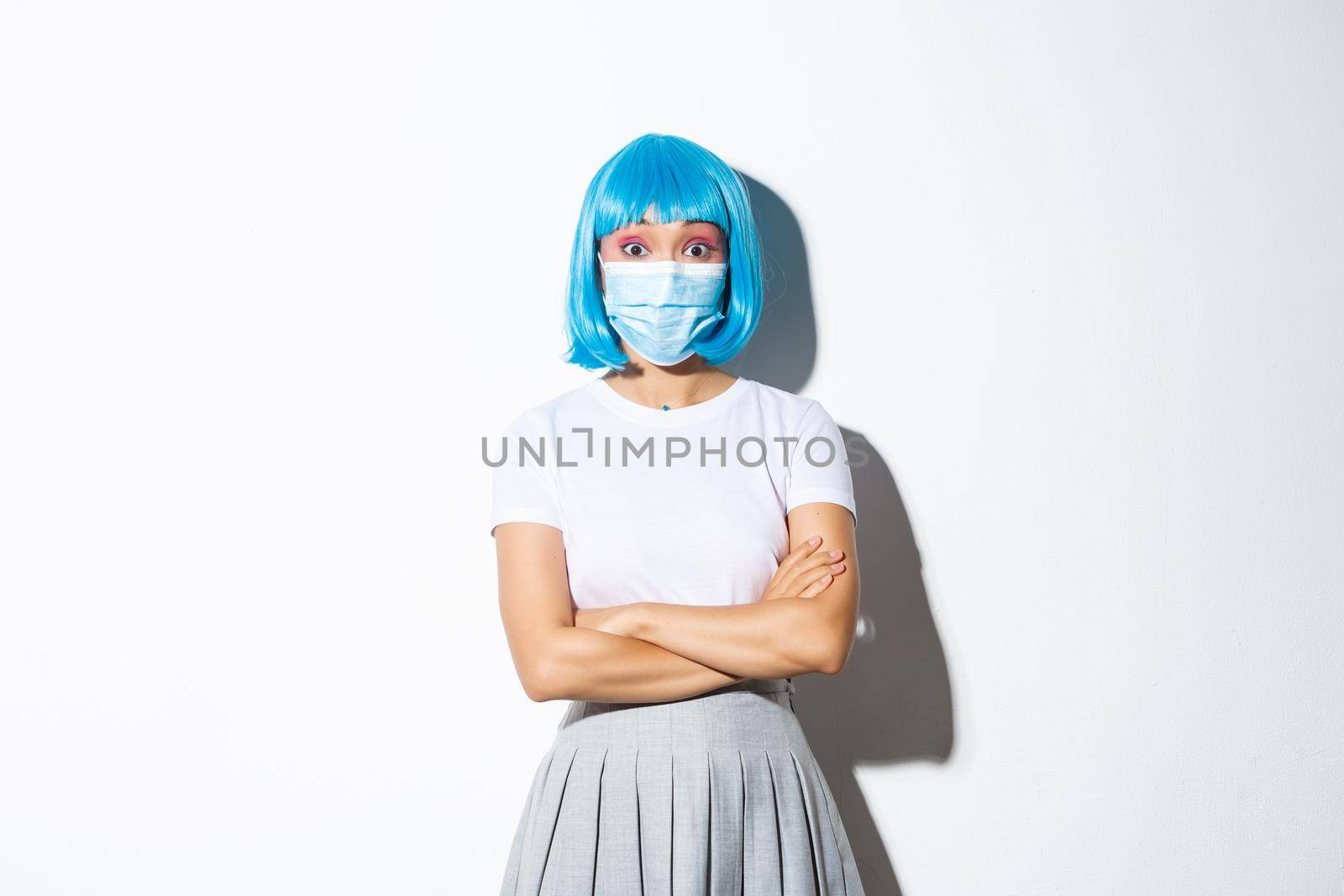 Concept of halloween celebration and covid-19 pandemic. Asian girl with blue wig and medical mask looking surprised, standing over white background.