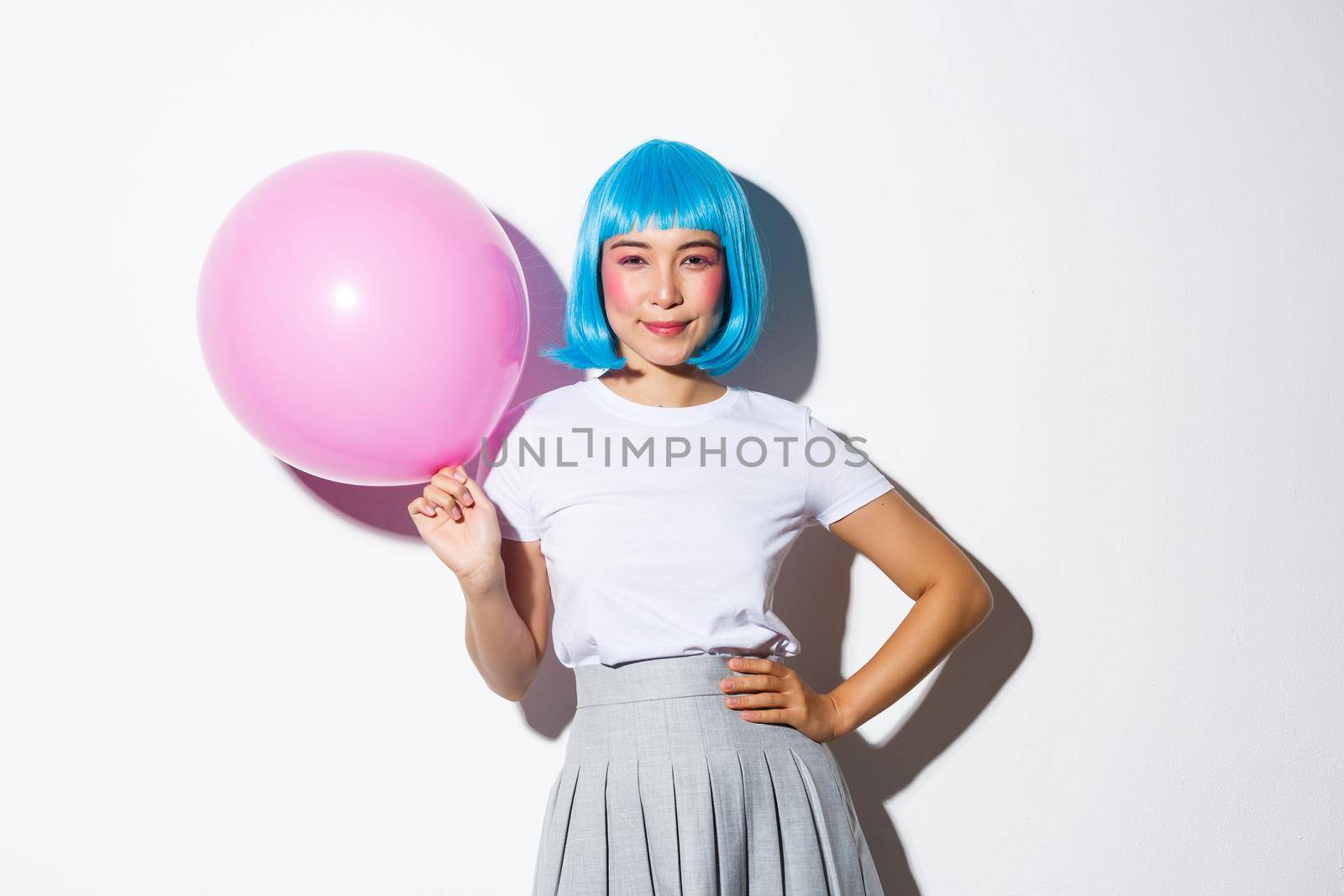 Image of coquettish sassy asian woman in blue wig, dressed-up for party, holding large pink balloon and smiling confident at camera.