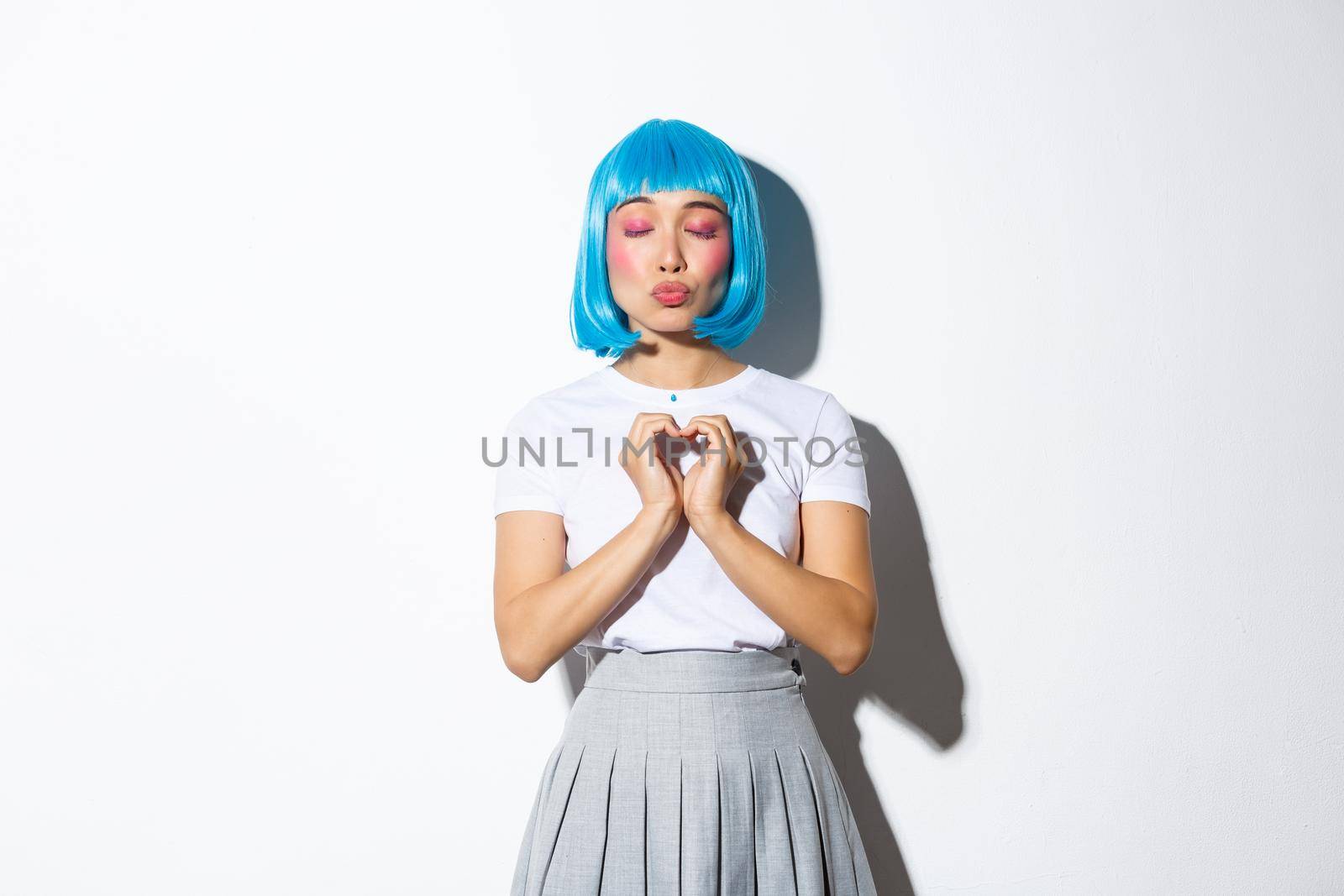Portrait of lovely asian girl in blue wig and halloween costume, close eyes and pouting while showing heart gesture, daydreaming, standing over white background.