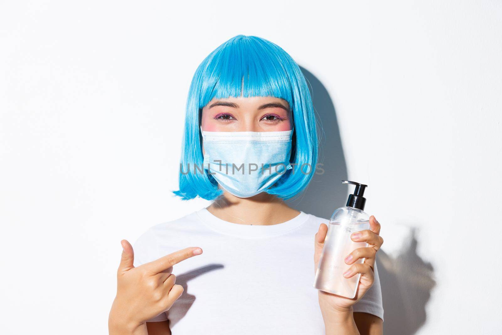 Concept of halloween celebration and coronavirus. Close-up of attractive asian woman in blue wig recommend using hand sanitizer, pointing at antiseptic, wearing medical mask.