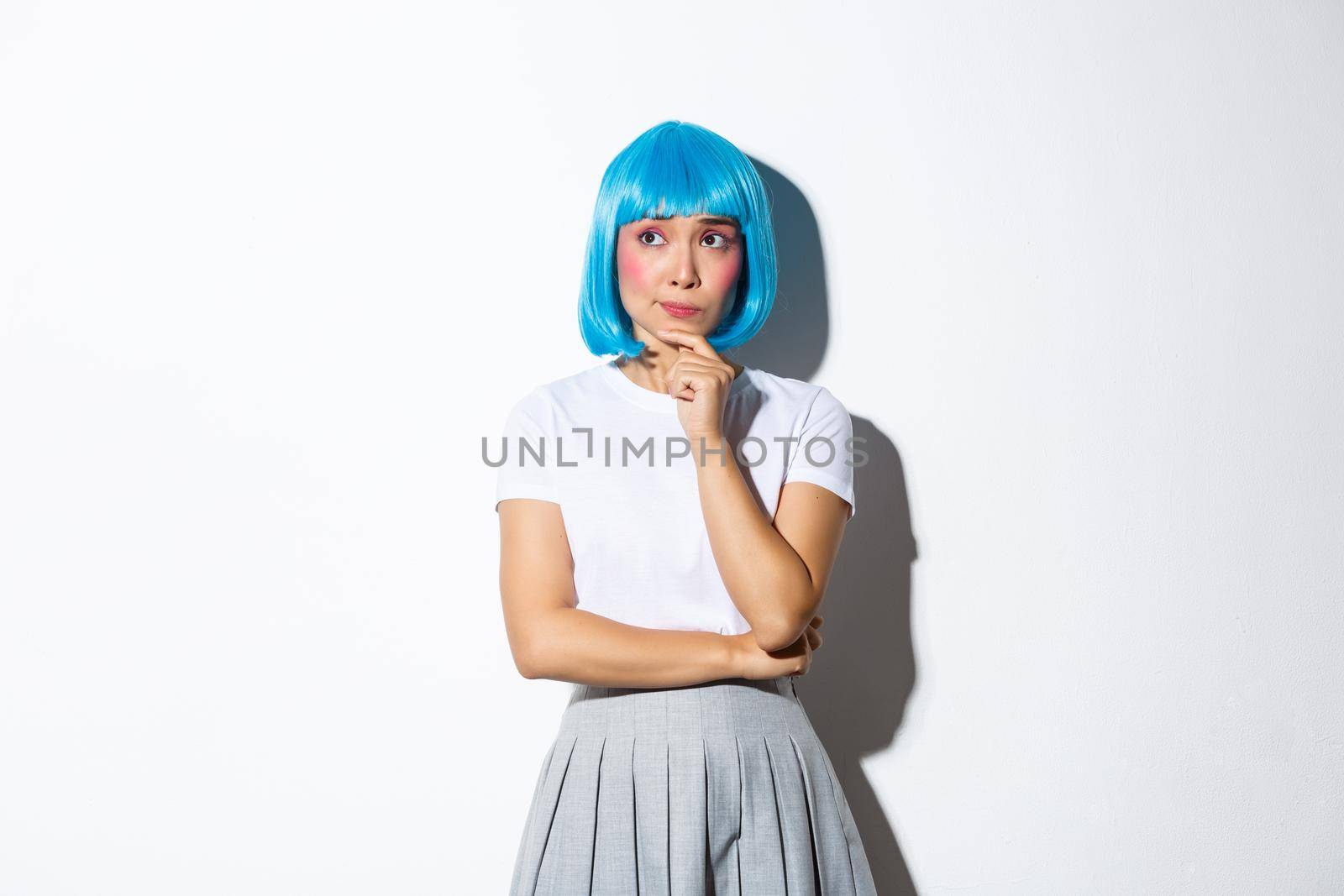 Image of troubled asian girl in blue short wig looking concerned and thoughtful, touching chin and gazing at upper left corner, thinking about something complicated, white background.