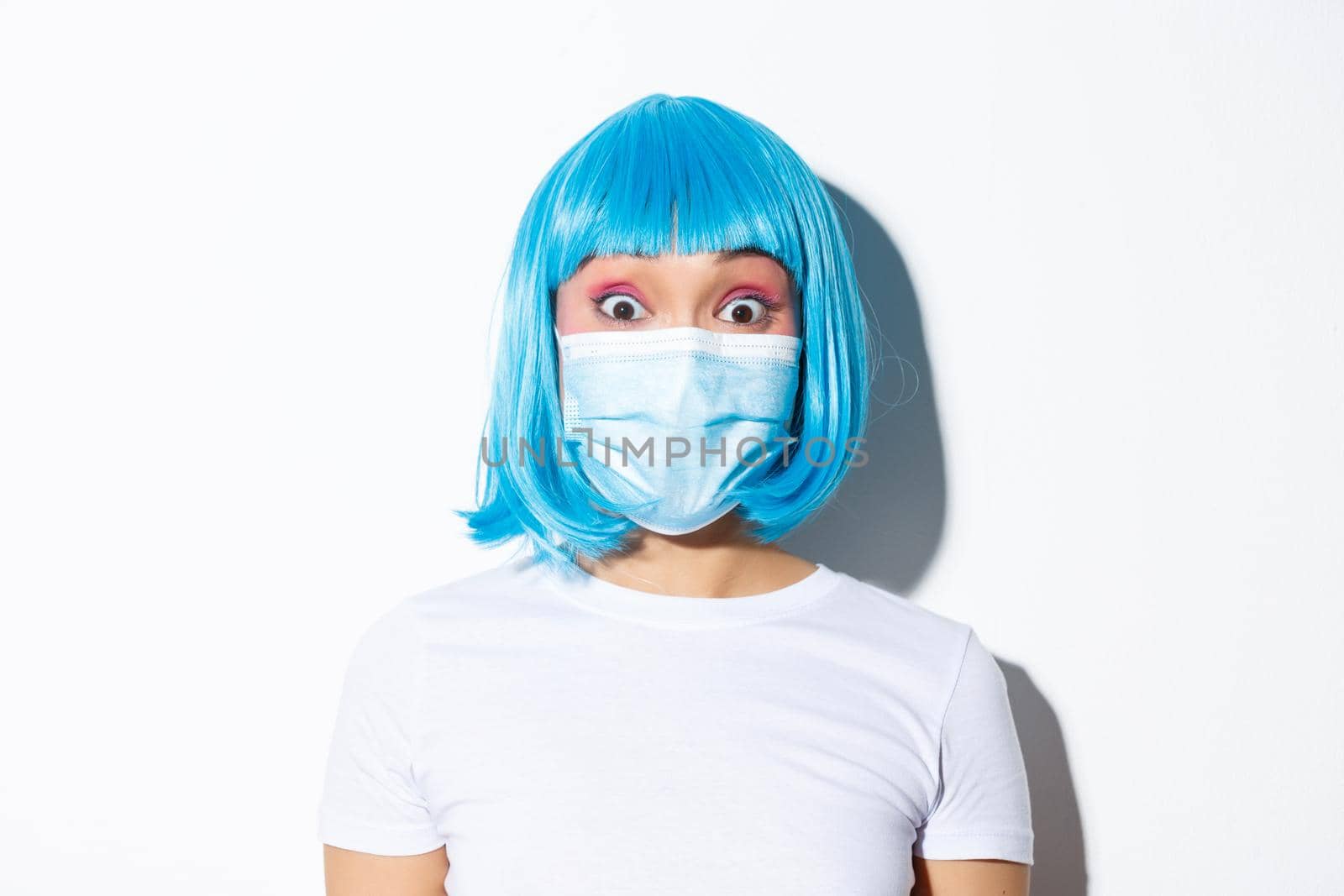Concept of halloween celebration and coronavirus. Close-up of surprised asian girl in medical mask and blue wig, looking at camera amazed, standing over white background.