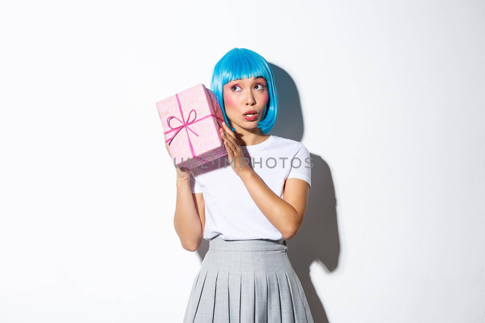 Cute asian girl in blue wig shaking box with gift, wonder whats inside, standing over white background.