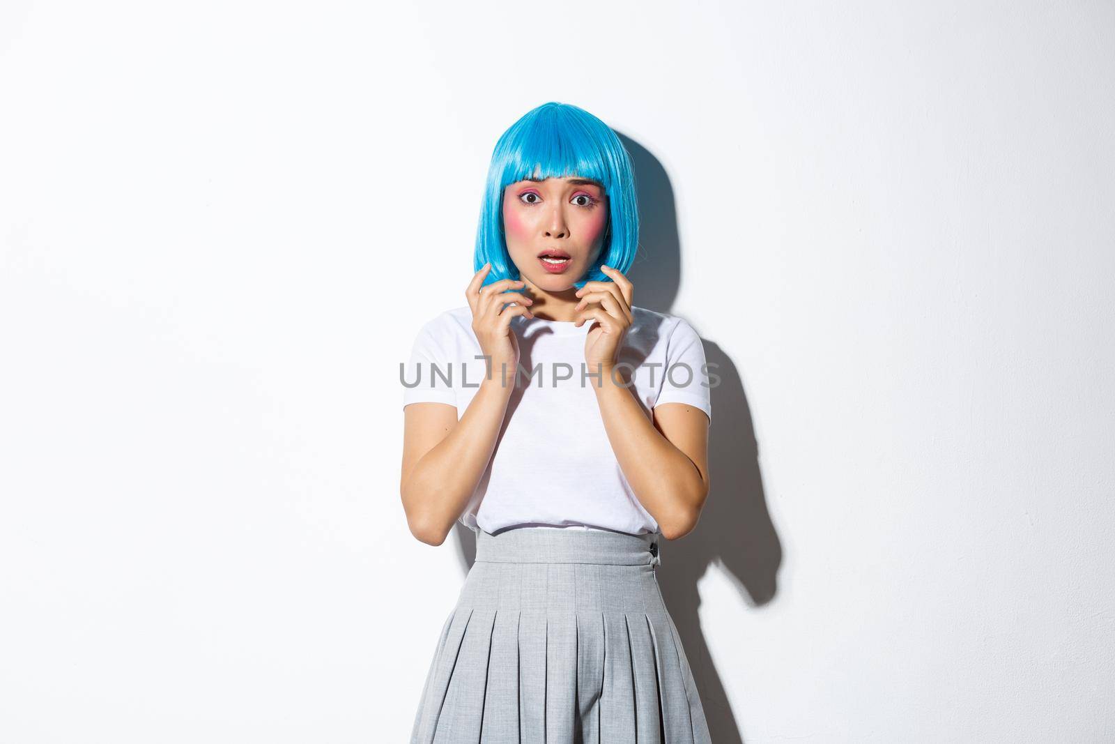 Portrait of scared asian woman in blue wig looking anxious and frightened, trembling from fear, wearing halloween costume, standing over white background.
