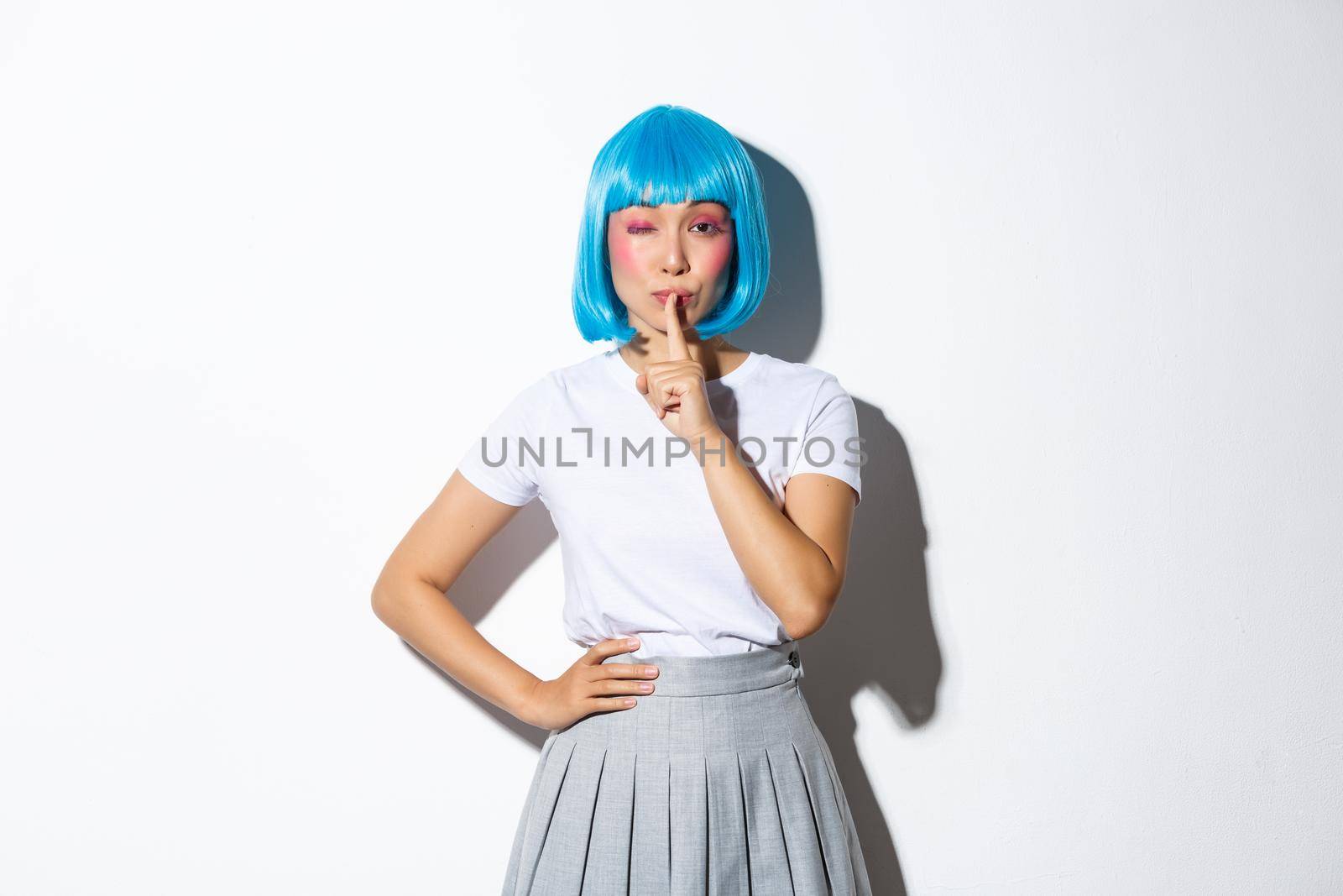 Sassy asian girl in blue wig share a secret, winking and shushing at camera with flirty expression, wearing blue short wig and halloween costume, white background.