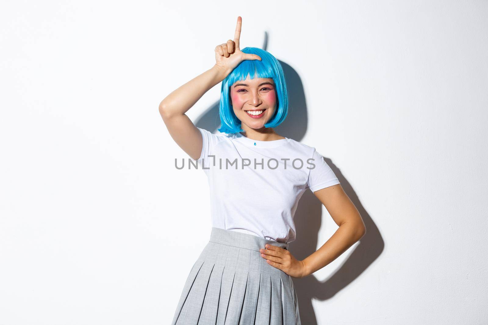 Portrait of beautiful silly asian girl celebrating halloween, showing loser gesture on forehead and smiling, mocking someone, standing over white background.