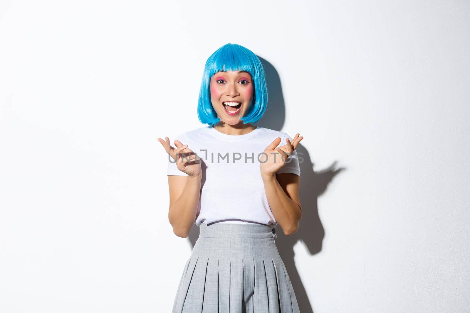 Portrait of happy and excited asian girl in blue short wig clap hands amused, smiling and looking at camera, standing over white background.