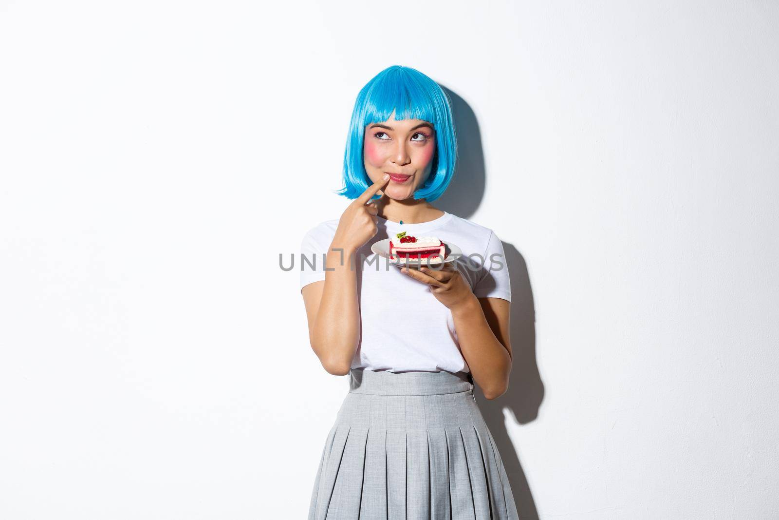 Coquettish beautiful asian girl in blue short wig, looking thoughtful and cunning at upper left corner, holding piece of cake, standing over white background.