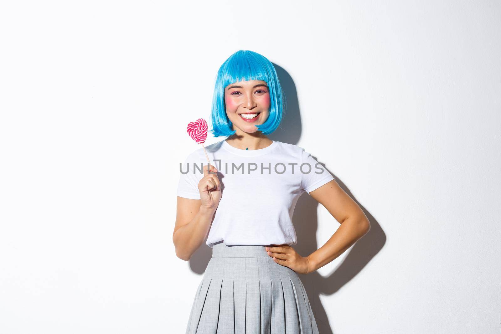 Image of cute asian girl in blue wig smiling happy, holding candy, going trick or treat on halloween in her schoolgirl costume, standing over white background.