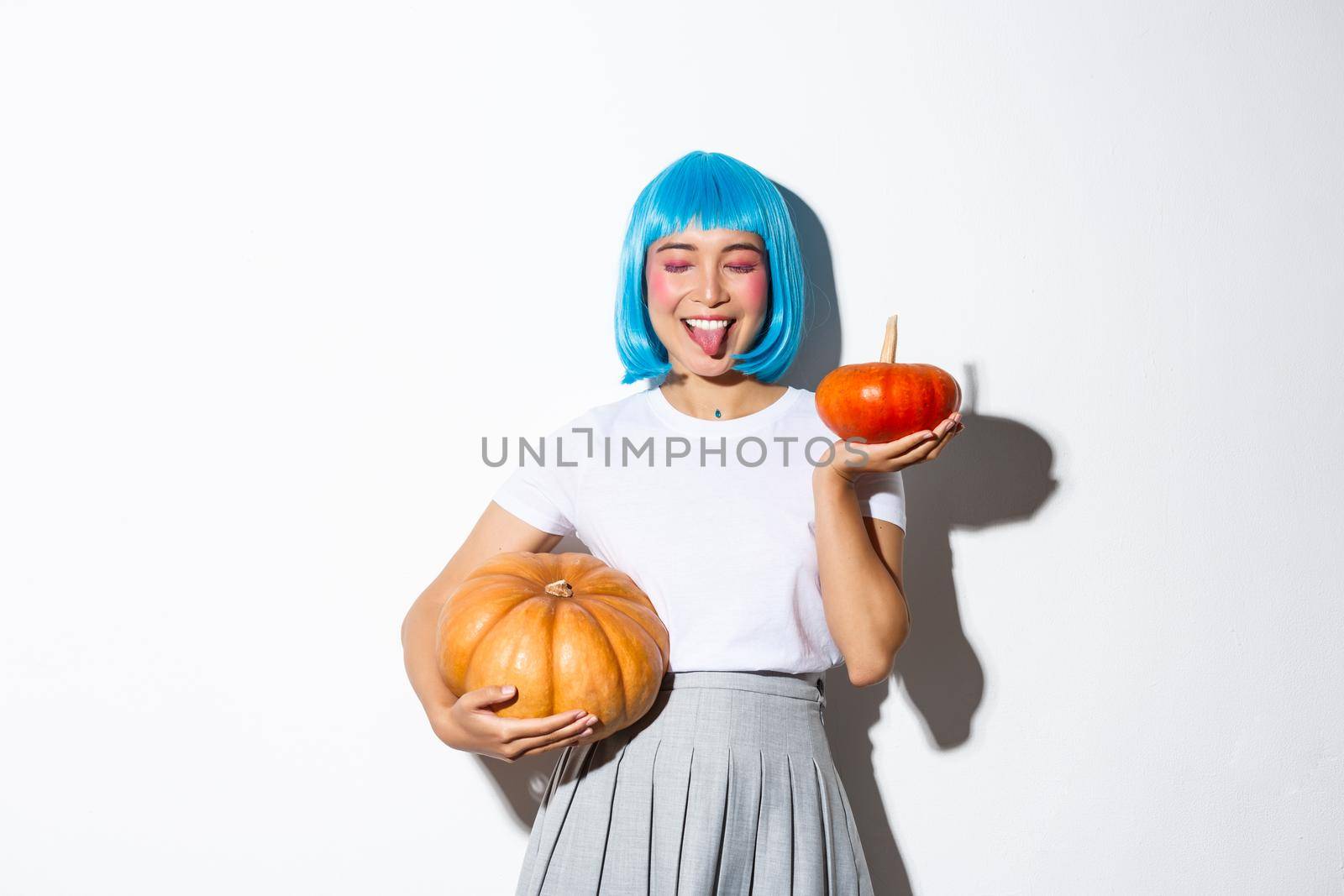 Happy attractive japanese girl in blue party wig, close eyes and showing tongue joyfully, celebrating halloween, holding two pumpkins.