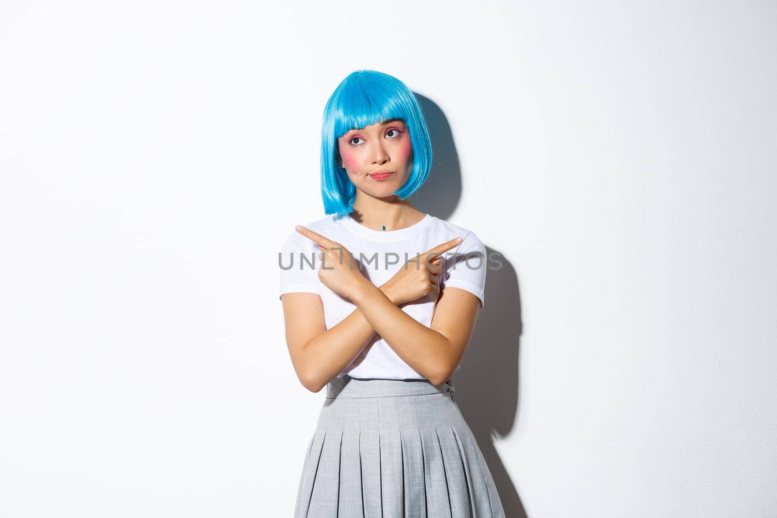 Indecisive beautiful asian girl in blue wig, pointing fingers sideways and looking puzzled while making choice, dressed for halloween, standing over white background.