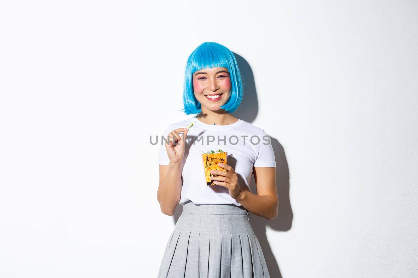 Portrait of cheerful asian girl in blue wig celebrating halloween, eating sweets from trick or treat bag, standing over white background.