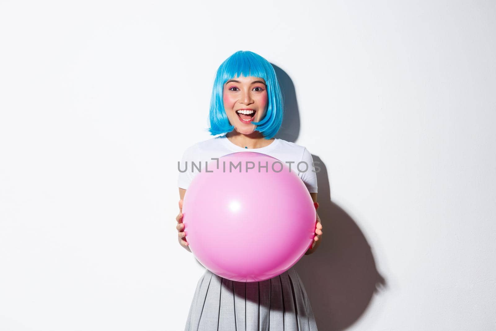 Image of cheerful asian girl in blue wig, celebrating holiday, wearing outfit for halloween party, holding large pink balloon and looking amazed.