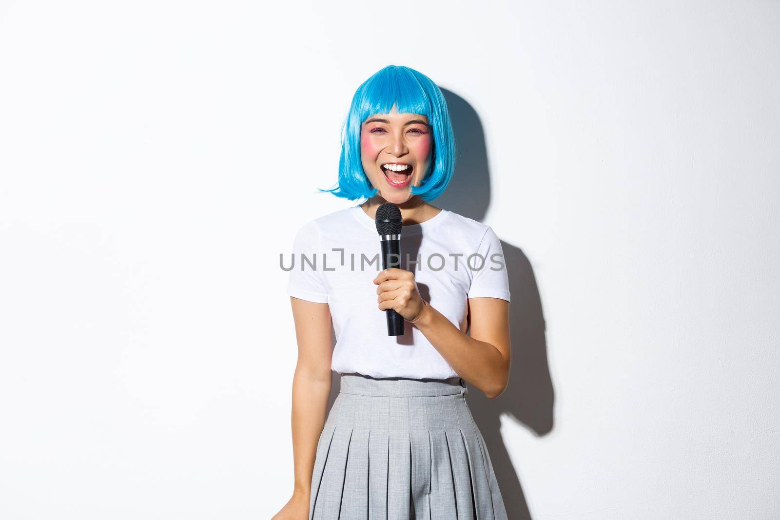 Image of beautiful japanese girl in blue anime wig, singing karaoke in microphone, smiling happy, standing over white background.