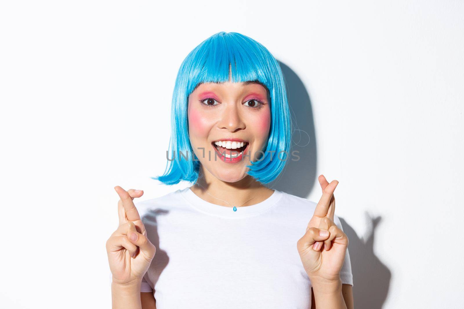 Close-up of hopeful cute asian girl in blue wig making wish, cross fingers good luck, standing over white background.