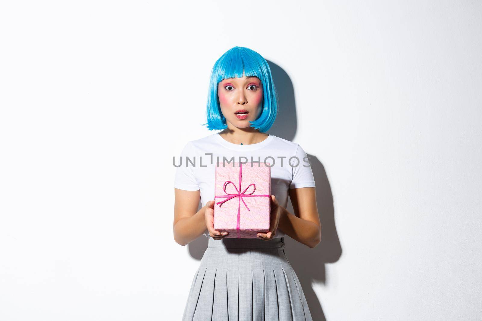Surprised asian girl in blue party wig looking at camera confused, holding giftbox, standing over white background.