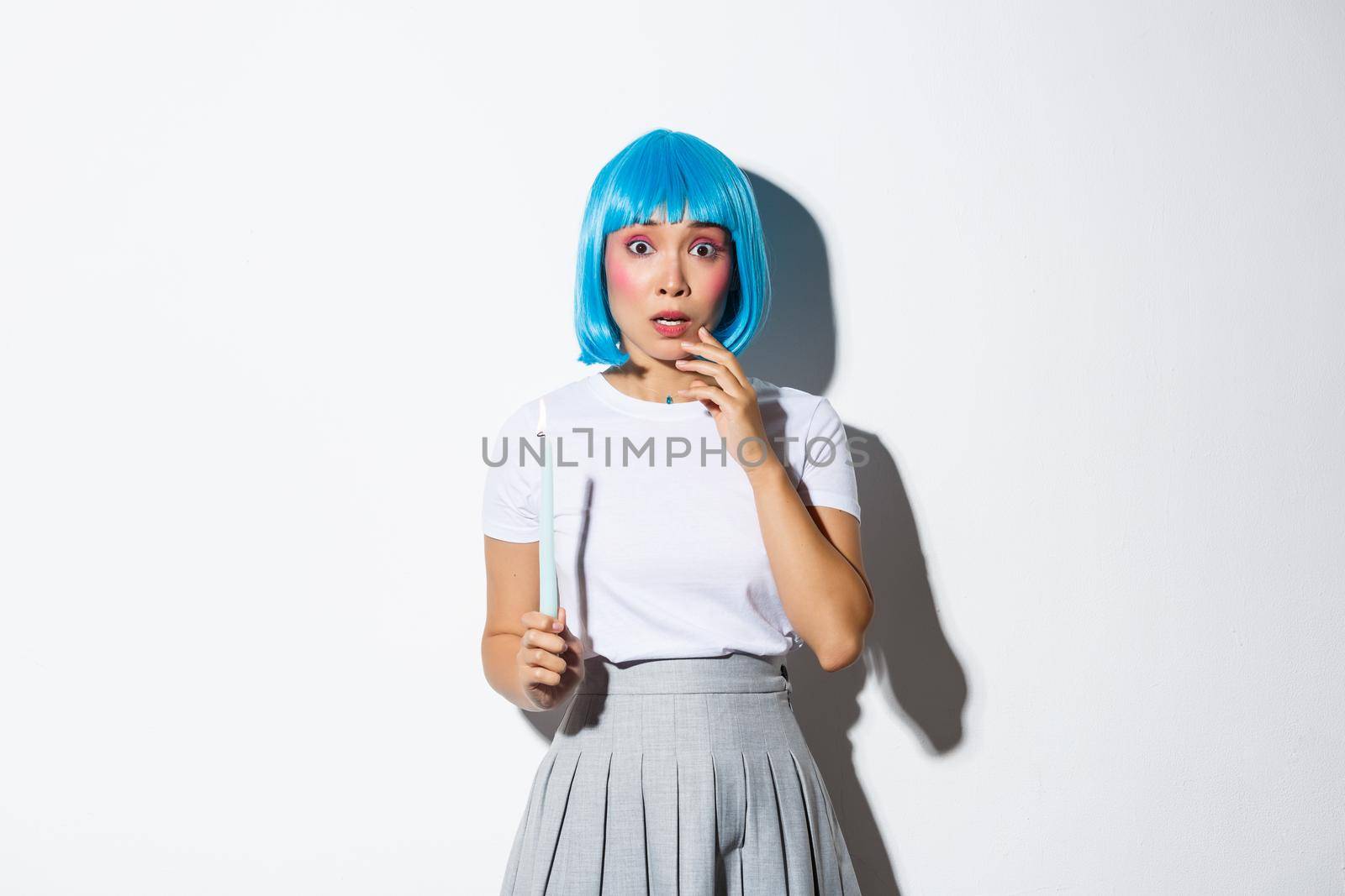 Concept of halloween. Image of scared timid asian girl in blue wig, holding candle and looking at something scary.