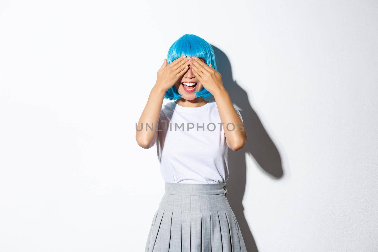 Cheerful party girl in blue wig close her eyes with hands and smiling, waiting for surprise, standing over white background.