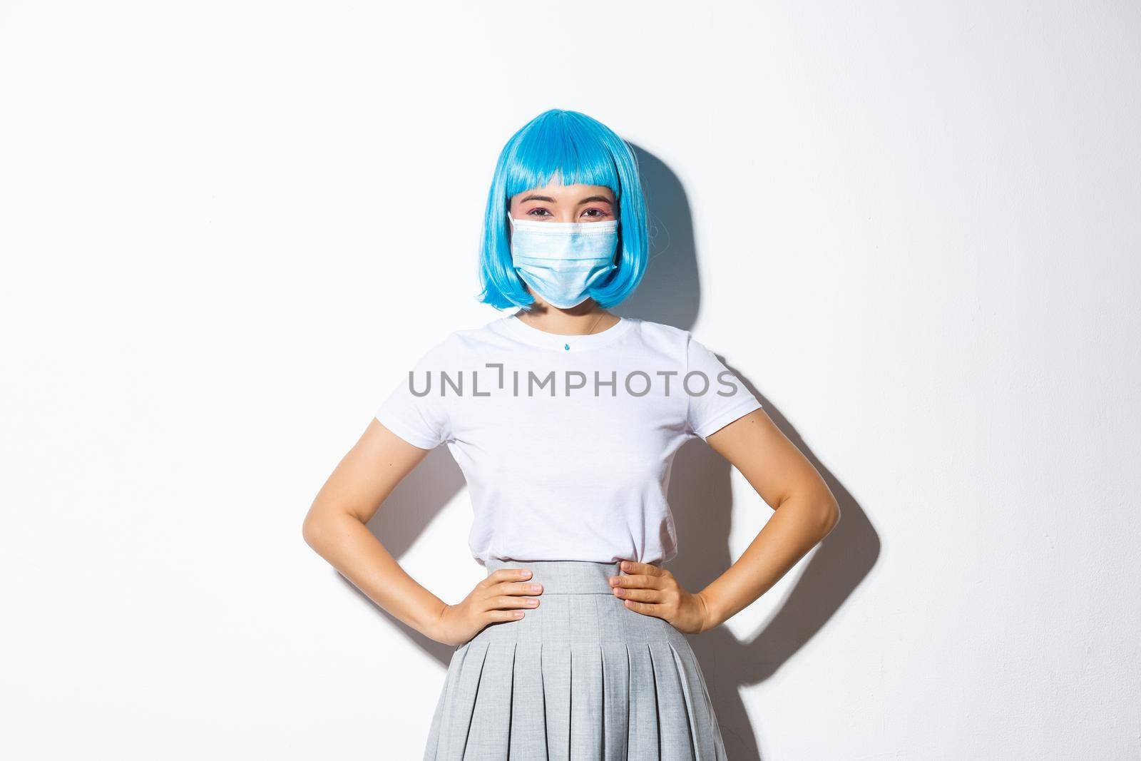 Image of cheerful asian girl ready for halloween party, protect herself from coronavirus as wearing medical mask, standing over white background in blue wig.