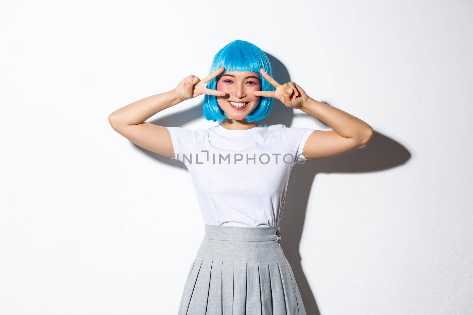 Joyful pretty asian woman in blue party wig, celebrating event, showing peace gestures and smiling happy, standing over white background.