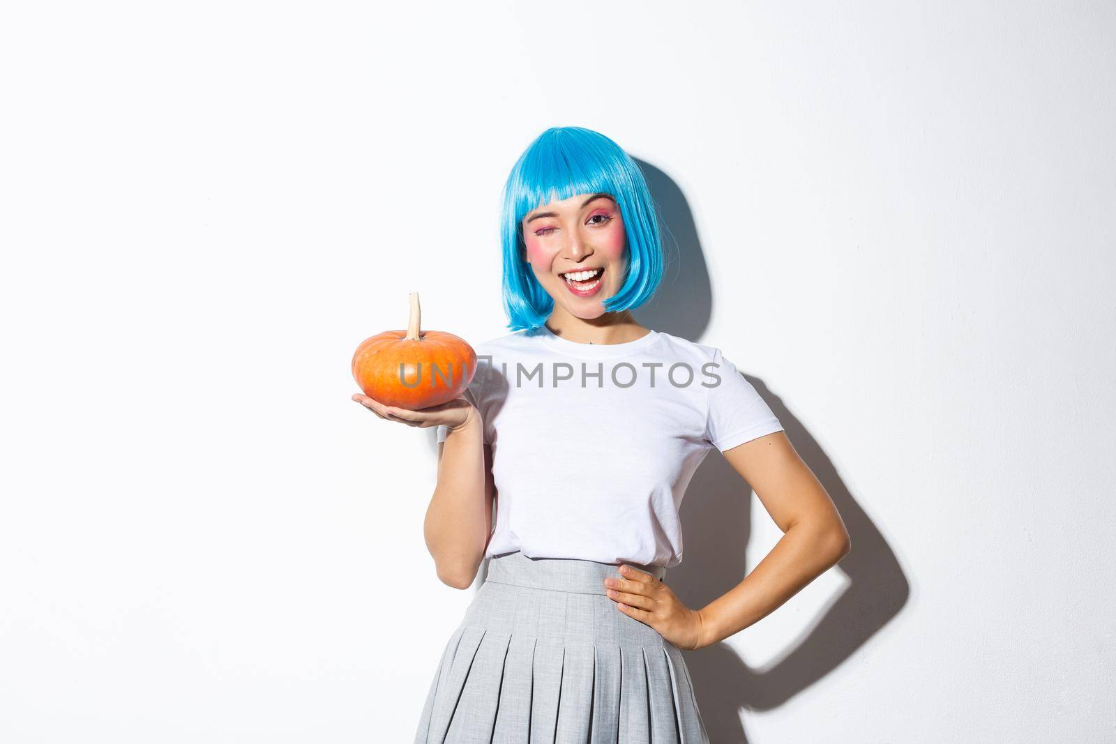 Portrait of coquettish asian girl in halloween costume and blue wig, holding cute pumpkin and winking at camera with happy expression, standing over white background.