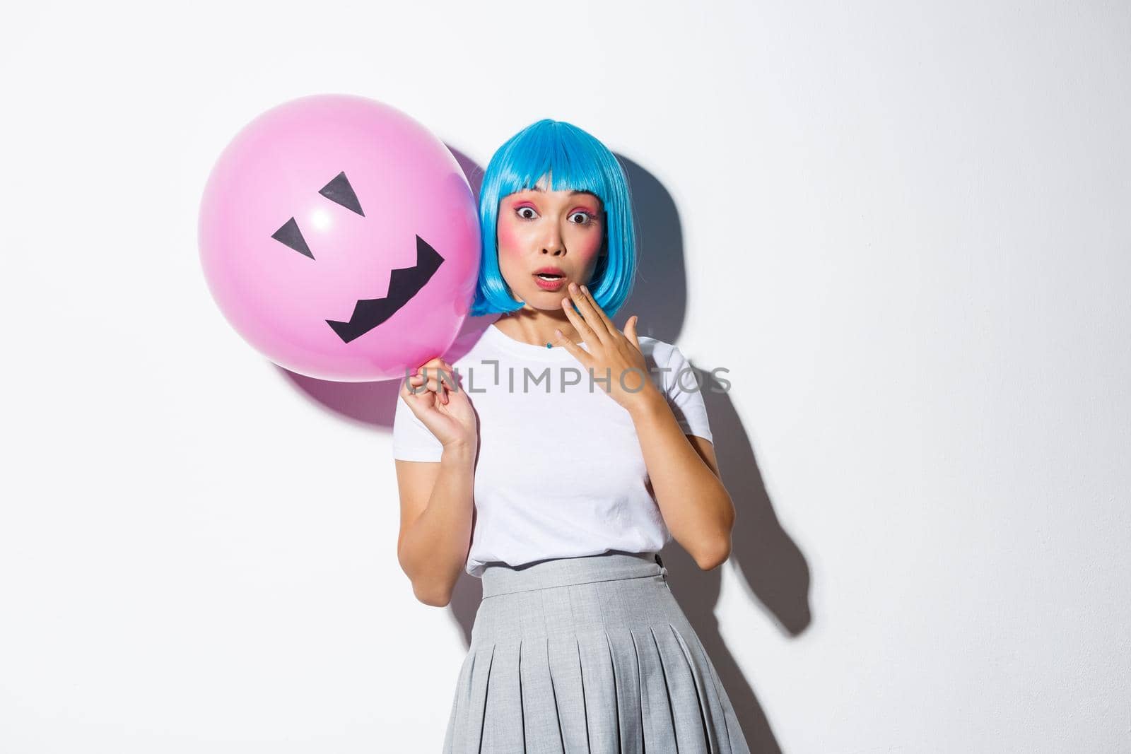 Image of cute asian girl in blue wig gasping surprised, holding pink balloon with scary face, standing over white background.