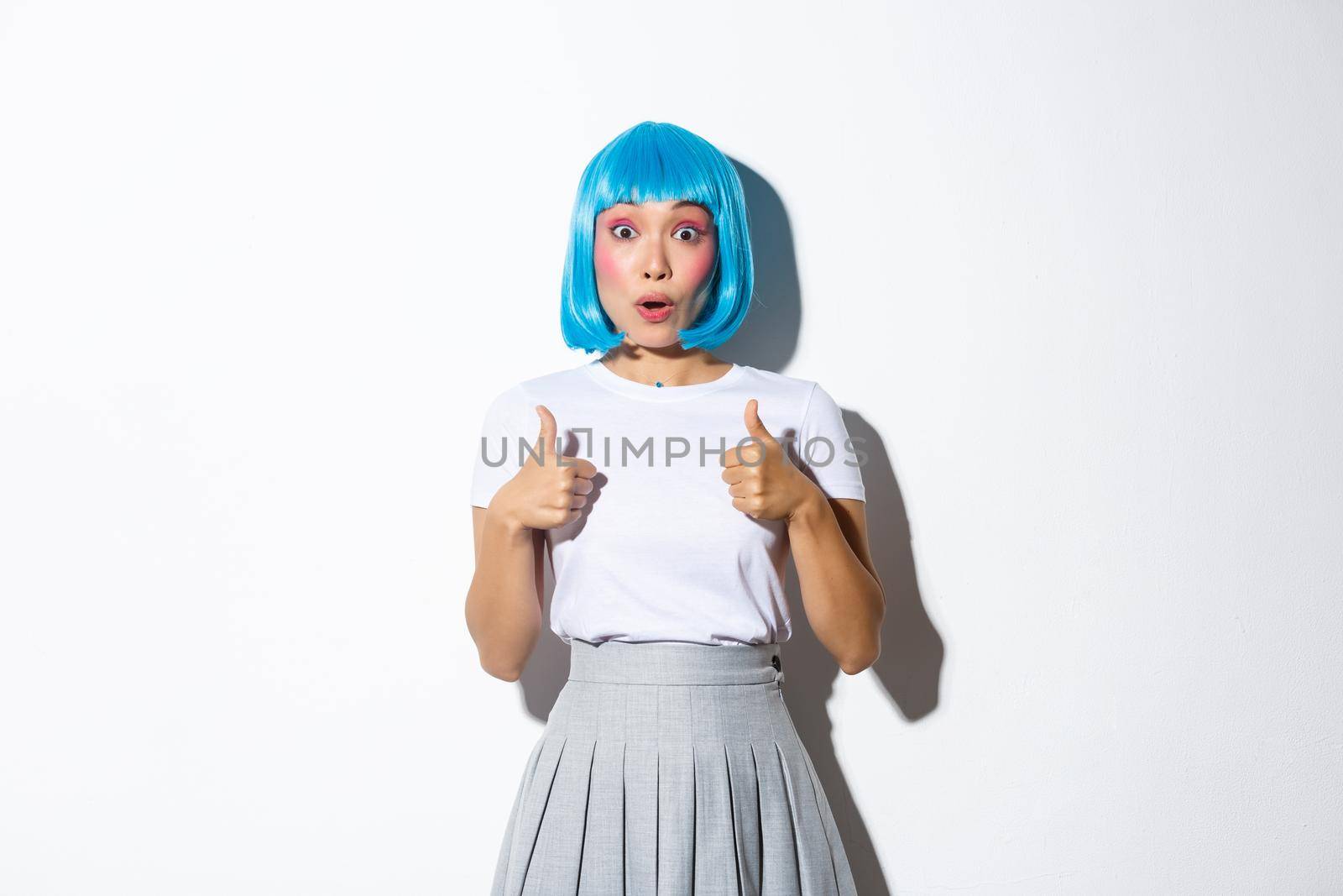 Surprised asian girl in blue wig gasping amazed, showing thumbs-up in approval, standing over white background.