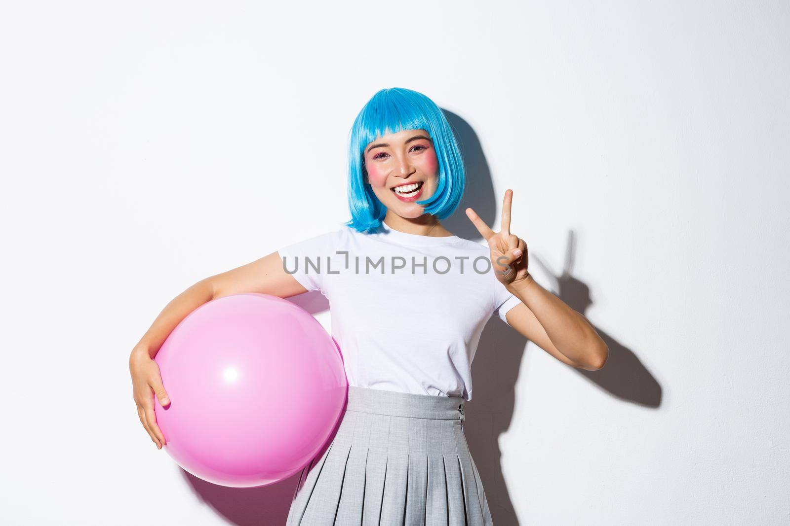 Image of cute asian girl in blue wig and halloween costume, showing peace gesture, holding large pink balloon.