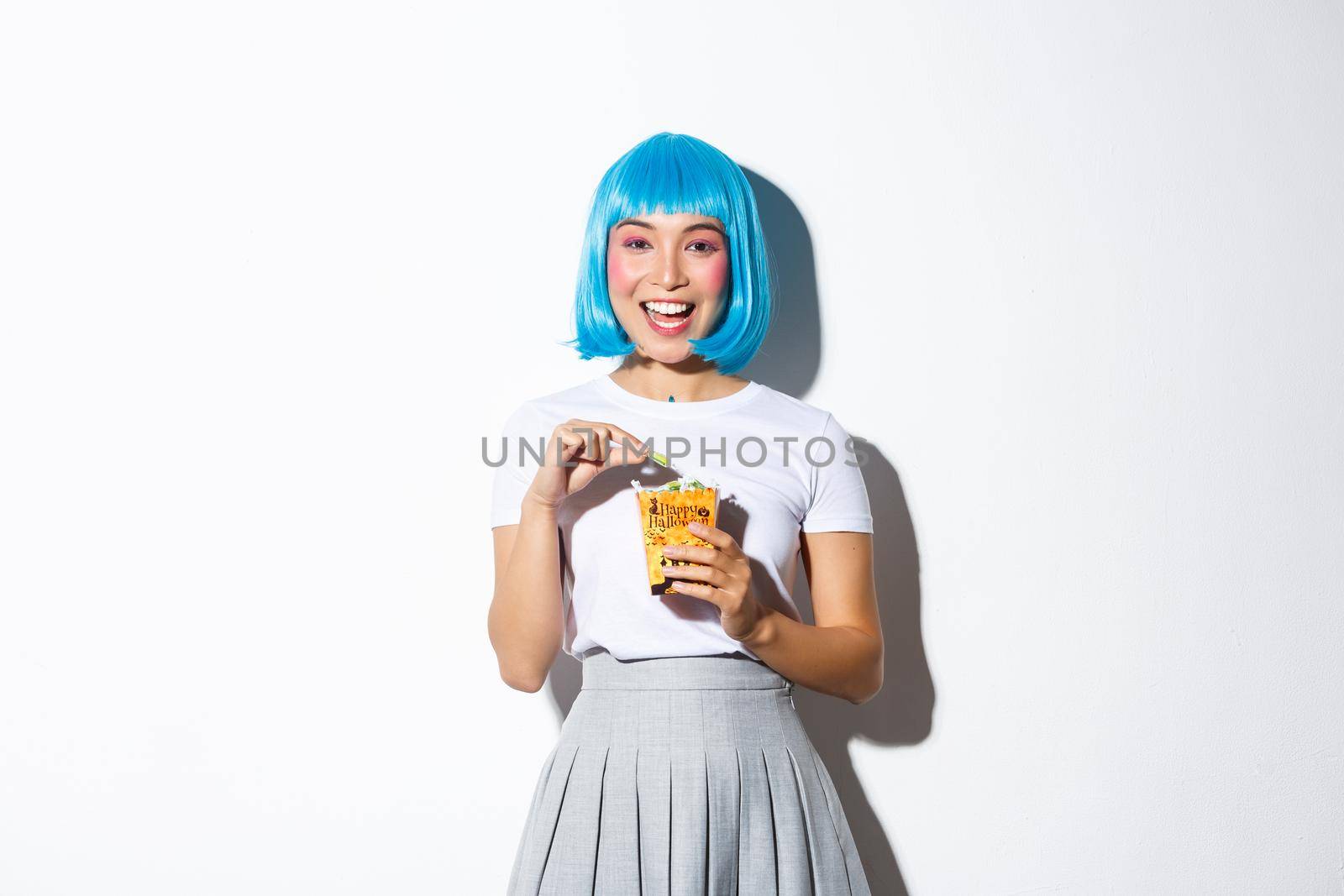 Image of cute asian girl celebrating halloween, eating sweets from trick or treat bag, wearing blue short wig and anime schoolgirl costume, standing over white background.