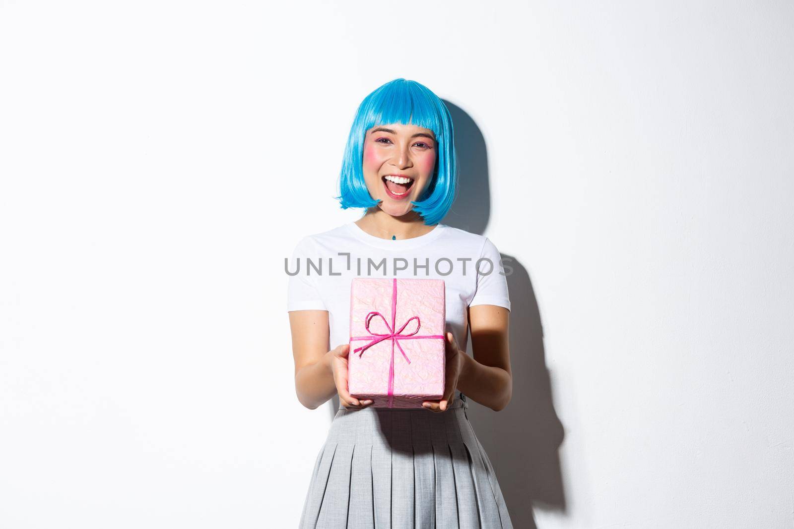 Image of cheerful cute asian girl in blue party wig, giving you a gift wrapped in pink paper, congratulating with something, standing over white background.