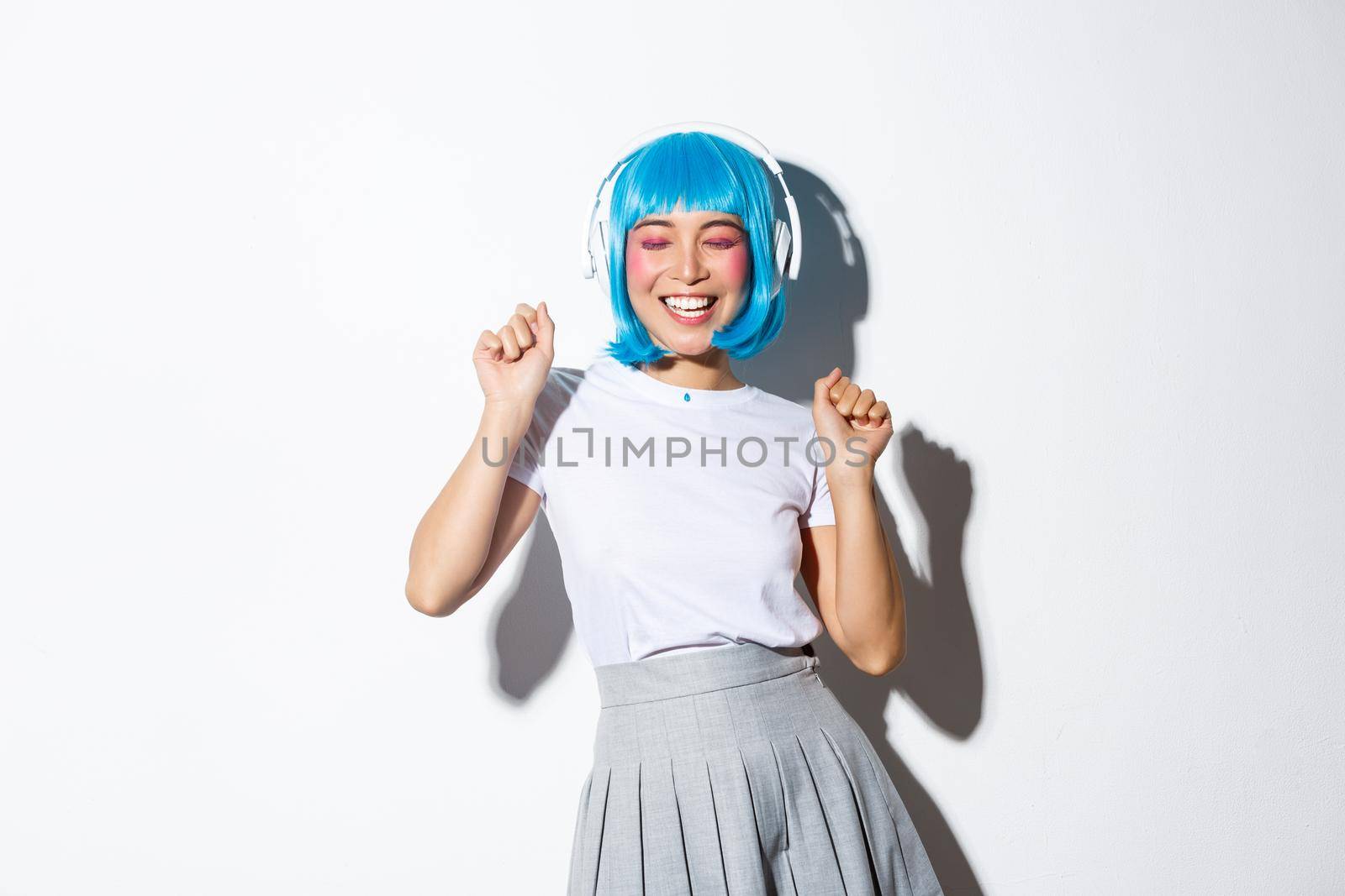 Portrait of happy asian girl in blue wig dancing, listening music in headphones over white background.