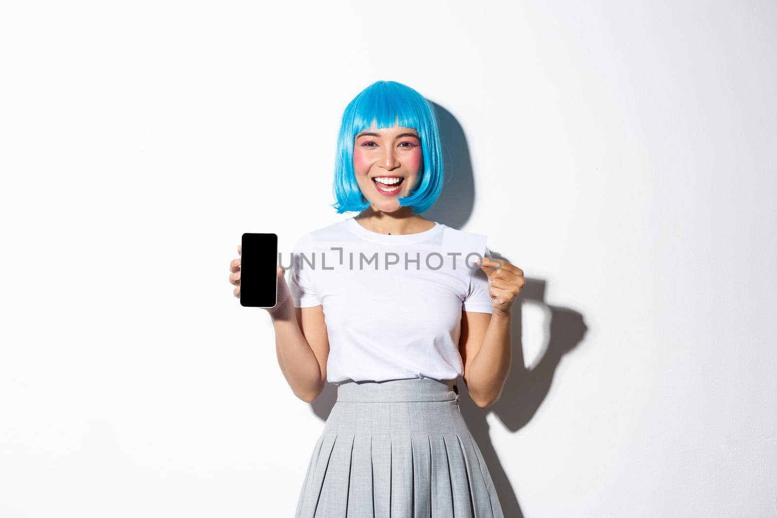 Happy pretty asian girl in blue short wig and party makeup, holding credit card and showing smartphone screen, looking happy, advertising application.