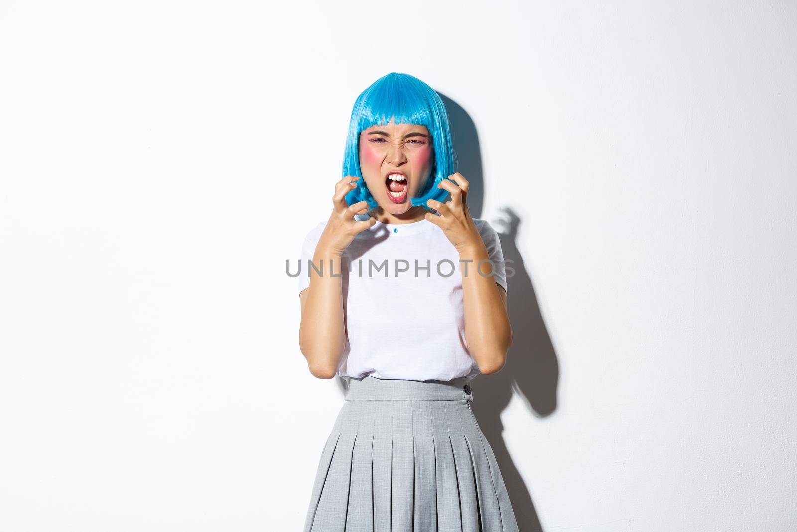 Portrait of angry asian girl in blue wig, yelling at someone with hatred, standing over white background.