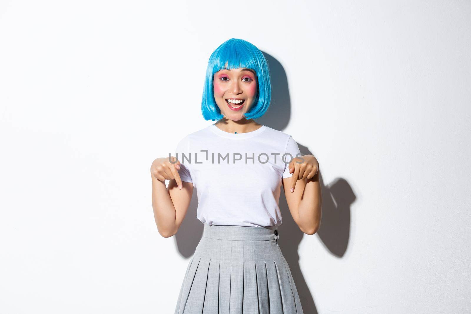 Image of happy silly asian girl showing you logo or product advertisement, pointing fingers down and smiling, wearing blue short wig, standing over white background.