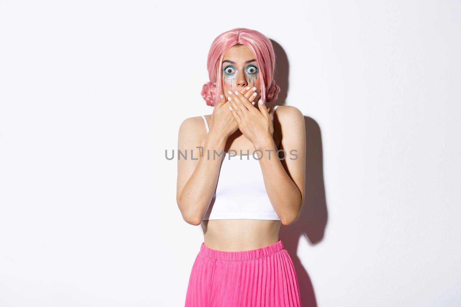 Image of shocked worried girl gasping, cover mouth with hands and looking scared, standing in halloween costume with pink wig and bright makeup.